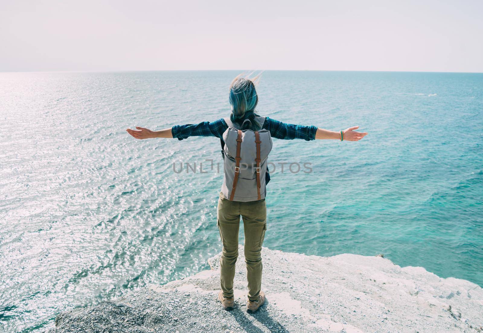 Happy explorer traveler young woman standing with raised arms on peak of rock and enjoying view of sea in summer, rear view