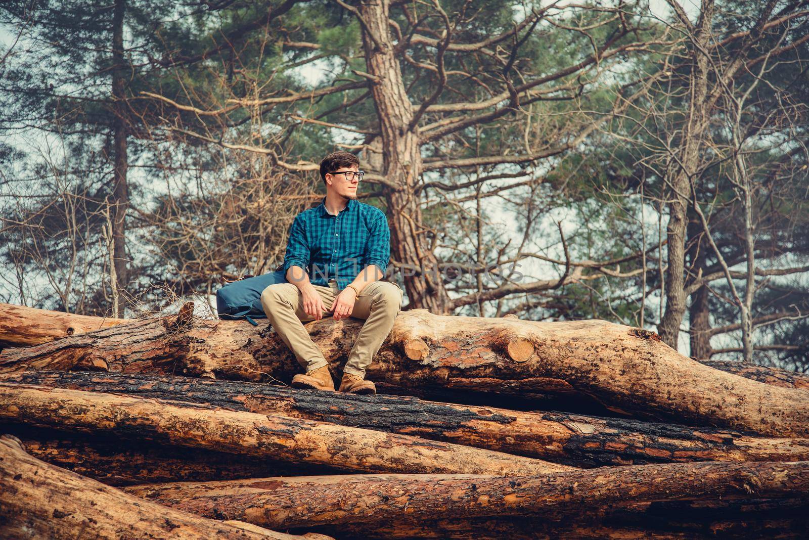 Explorer traveler young man with backpack sitting on felled tree trunk in the pine forest outdoor in summer