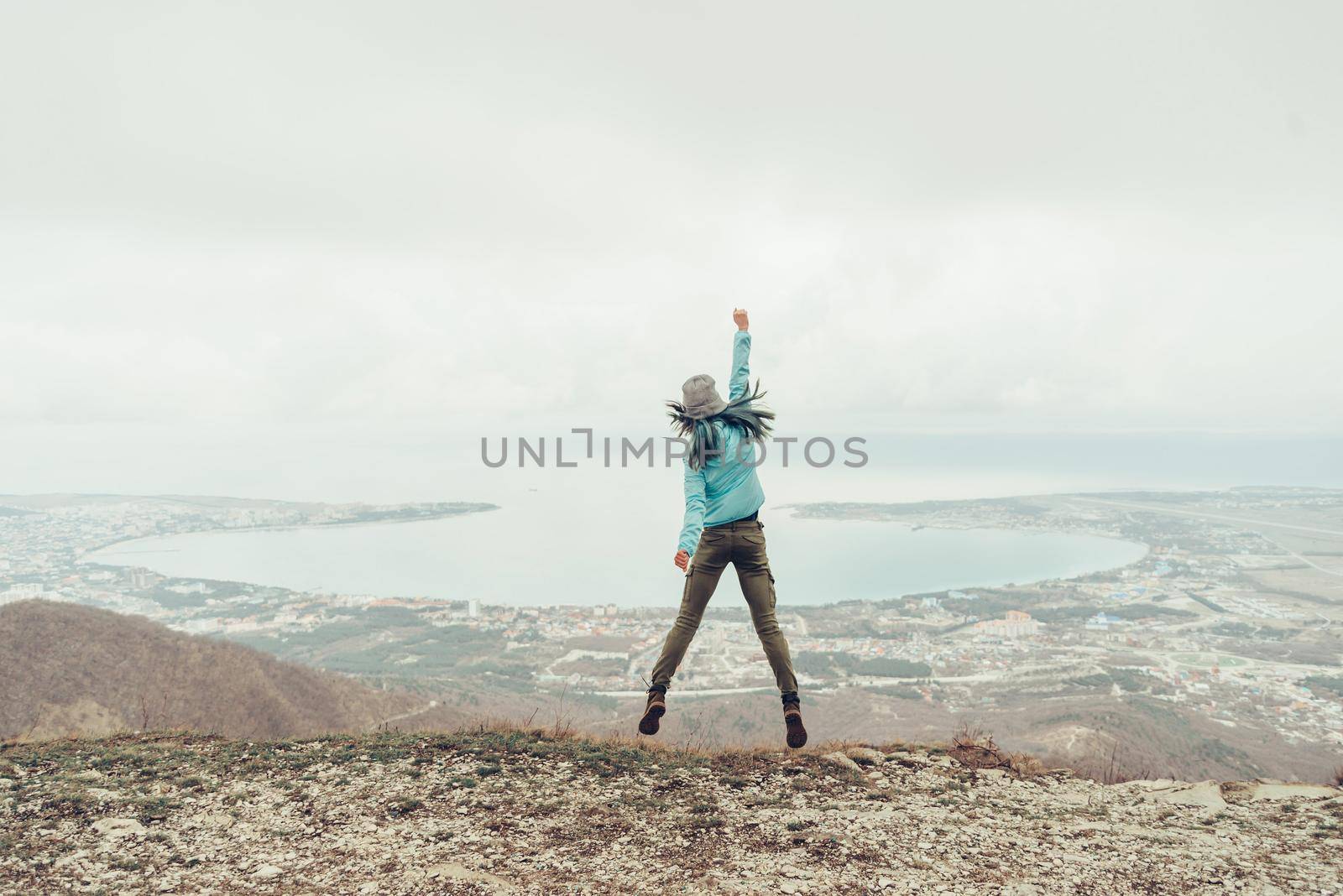 Happy girl jumping on peak of mountain above the sea bay, rear view