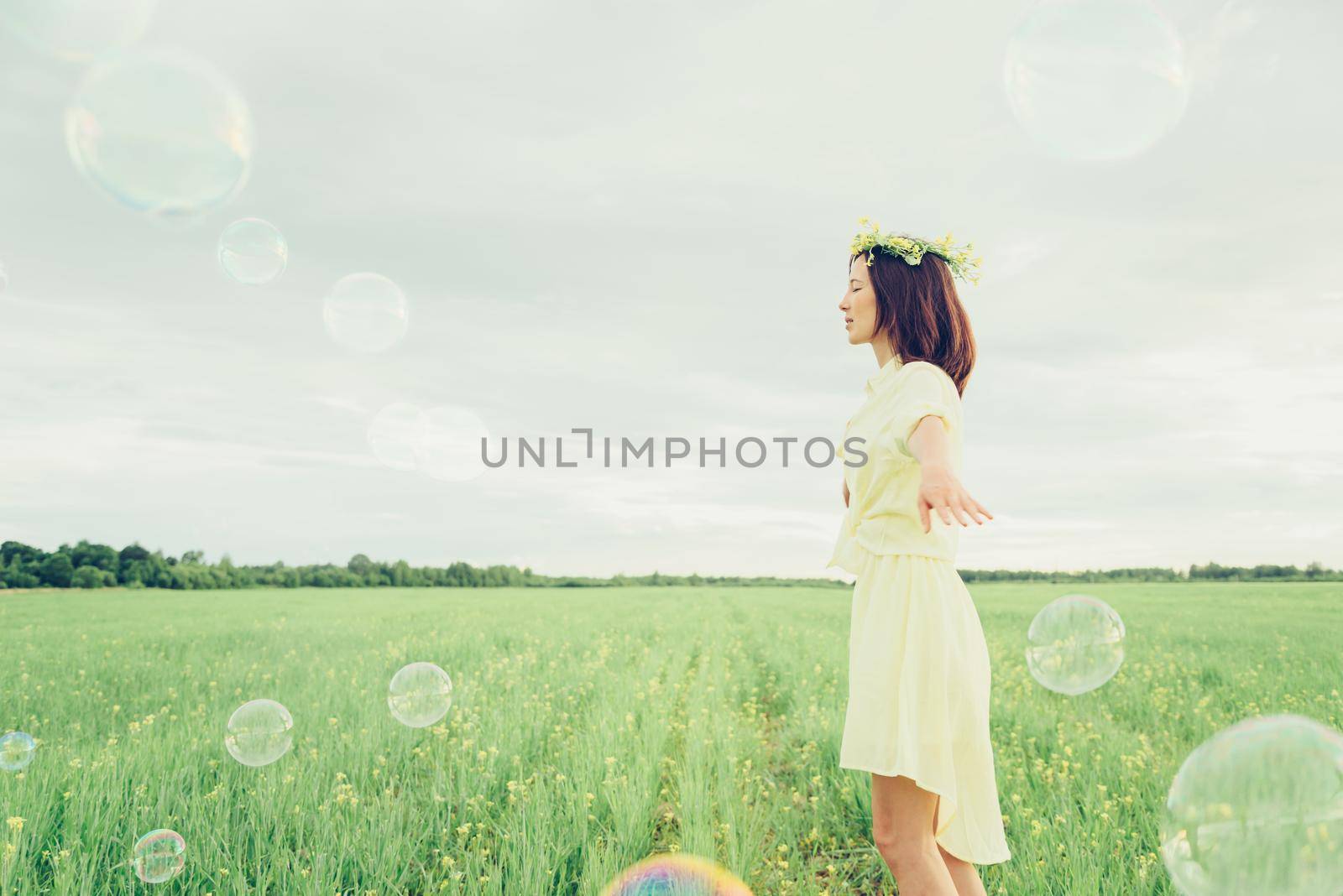 Happy beautiful young woman in flower wreath walking on summer meadow among soap bubbles