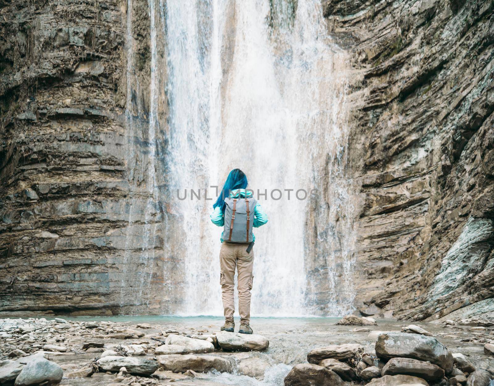 Traveler young woman with backpack standing on stone and enjoying view of beautiful waterfall outdoor, rear view
