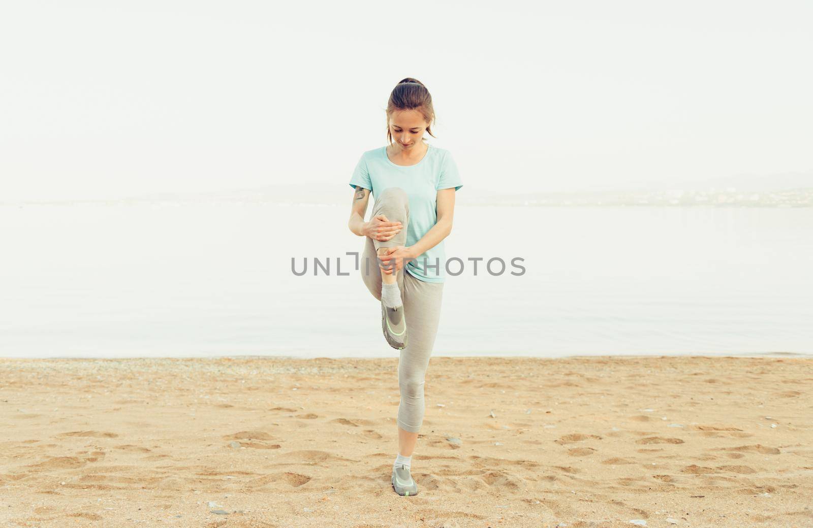 Sporty young woman stretching her legs and preparing to run on sand beach in summer, workout. Concept of healthy lifestyle