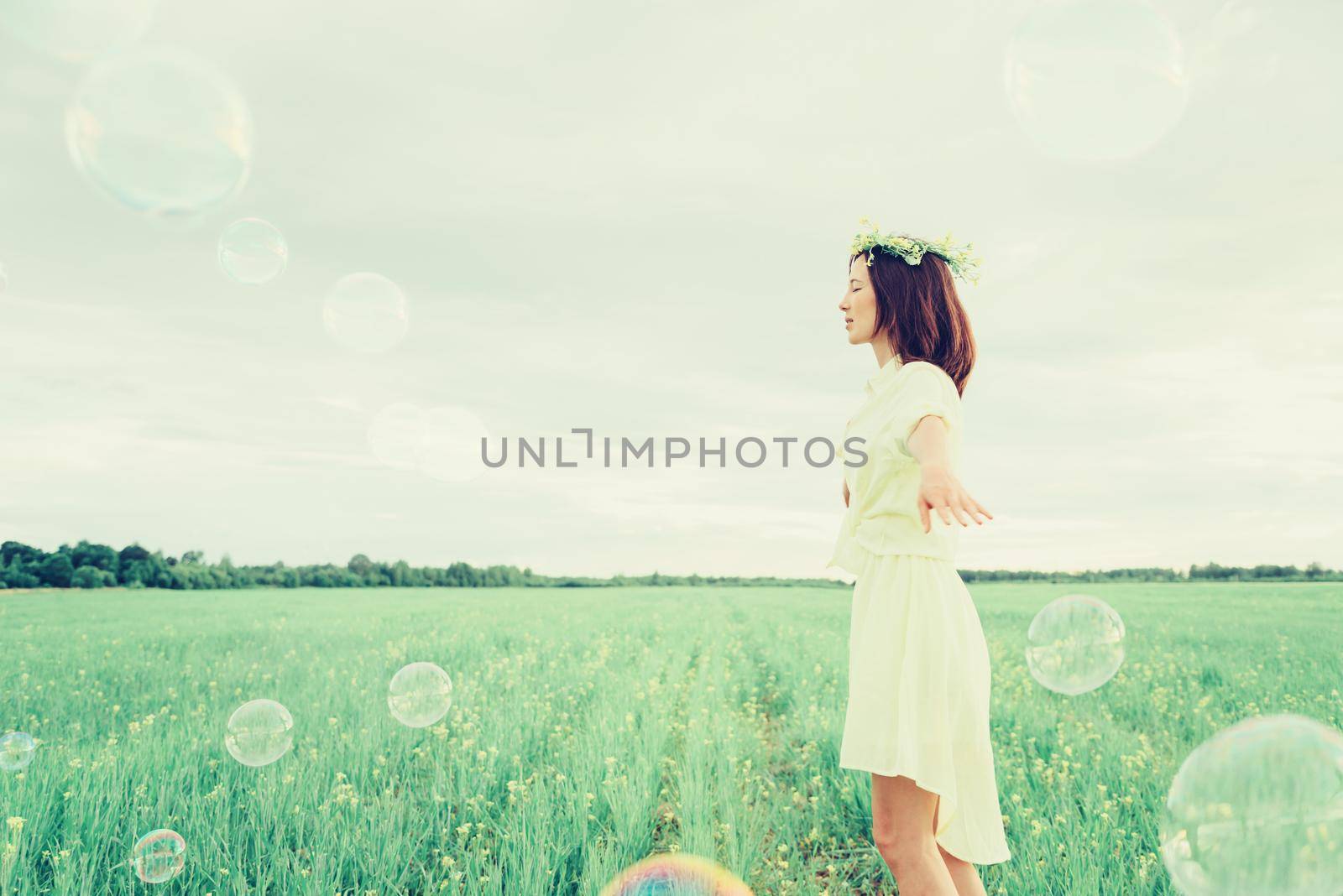 Happy beautiful girl in flower wreath walking on summer meadow among soap bubbles
