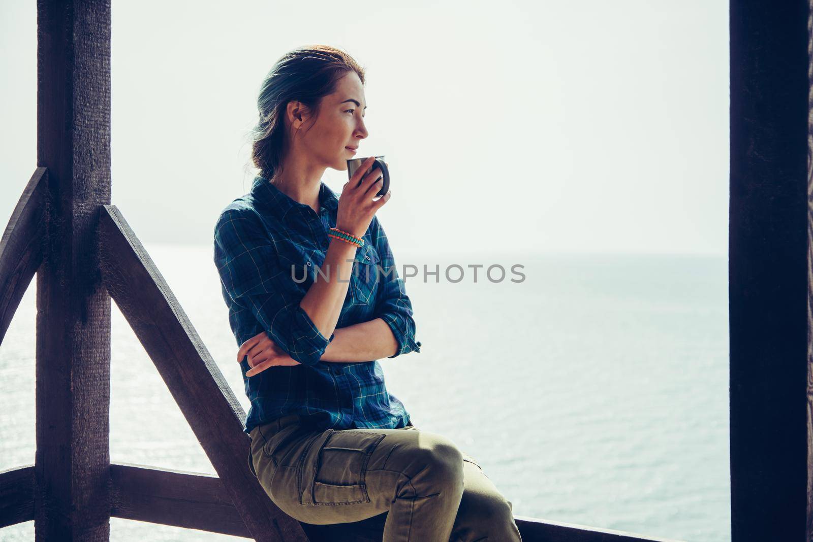 Smiling young woman sitting with cup in wooden arbor and enjoying view of sea