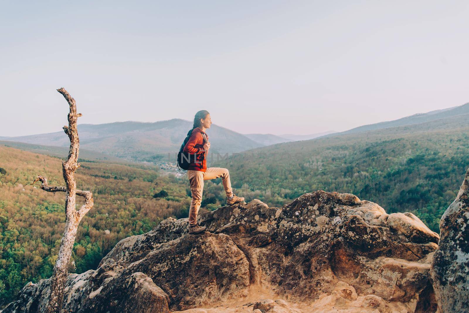Hiker young woman with backpack standing on peak of rock on background of mountains and looking into the distance in summer outdoor