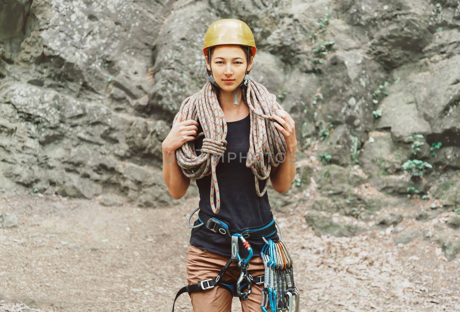 Young woman wearing in safety harness with climber equipment and helmet holding rope and looking at camera on background of rock