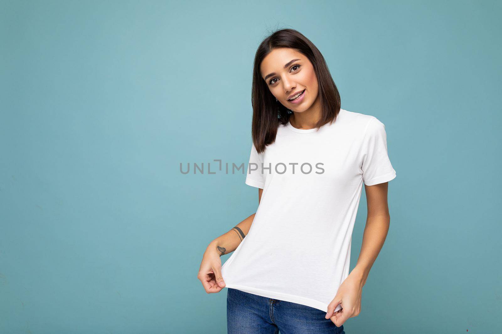 Portrait of positive cheerful fashionable smiling young brunette woman in casual white t-shirt for mockup isolated on blue background with copy space by TRMK