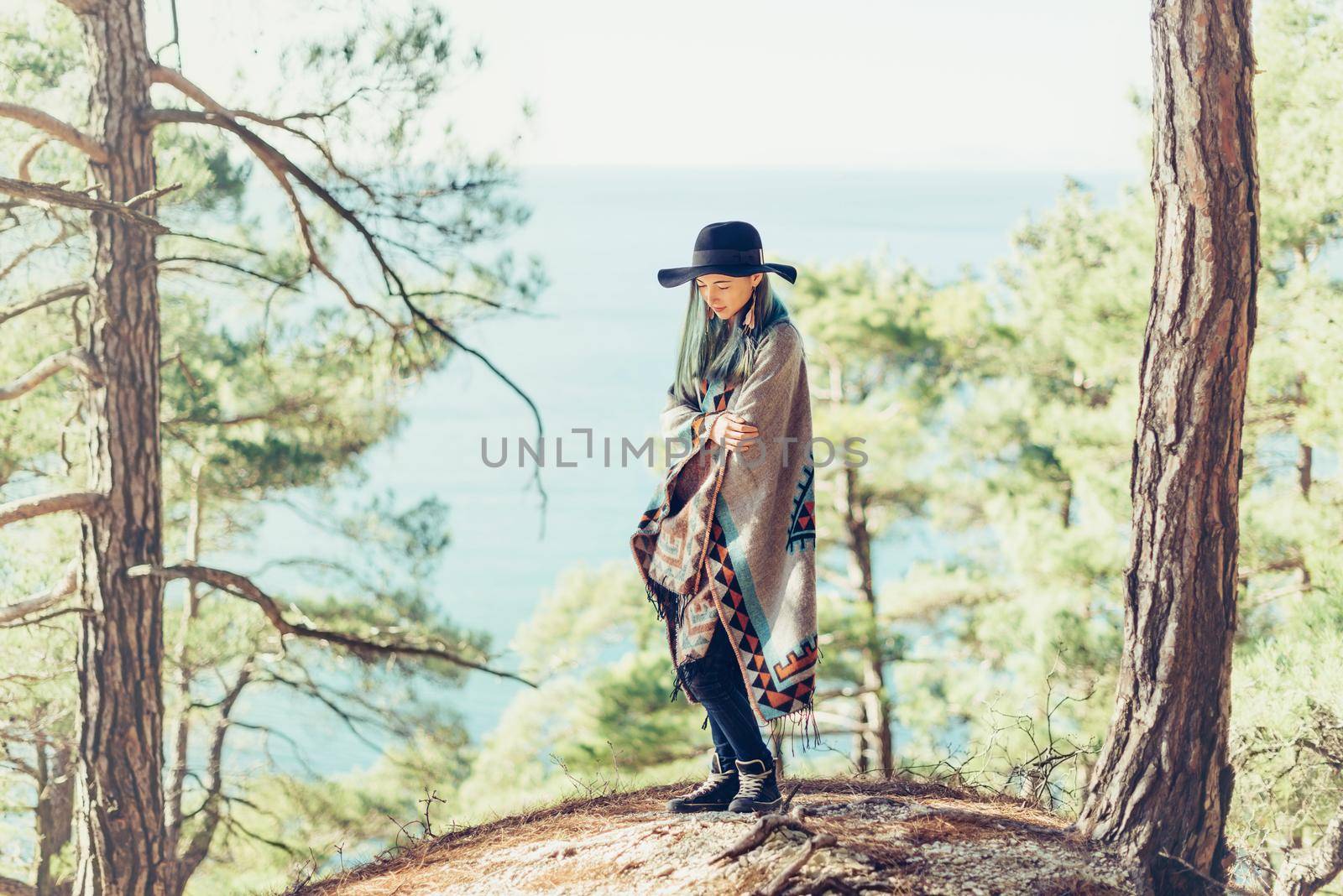 Fashionable young woman wearing in hat and poncho standing on coast in the forest among pine trees on background of sea