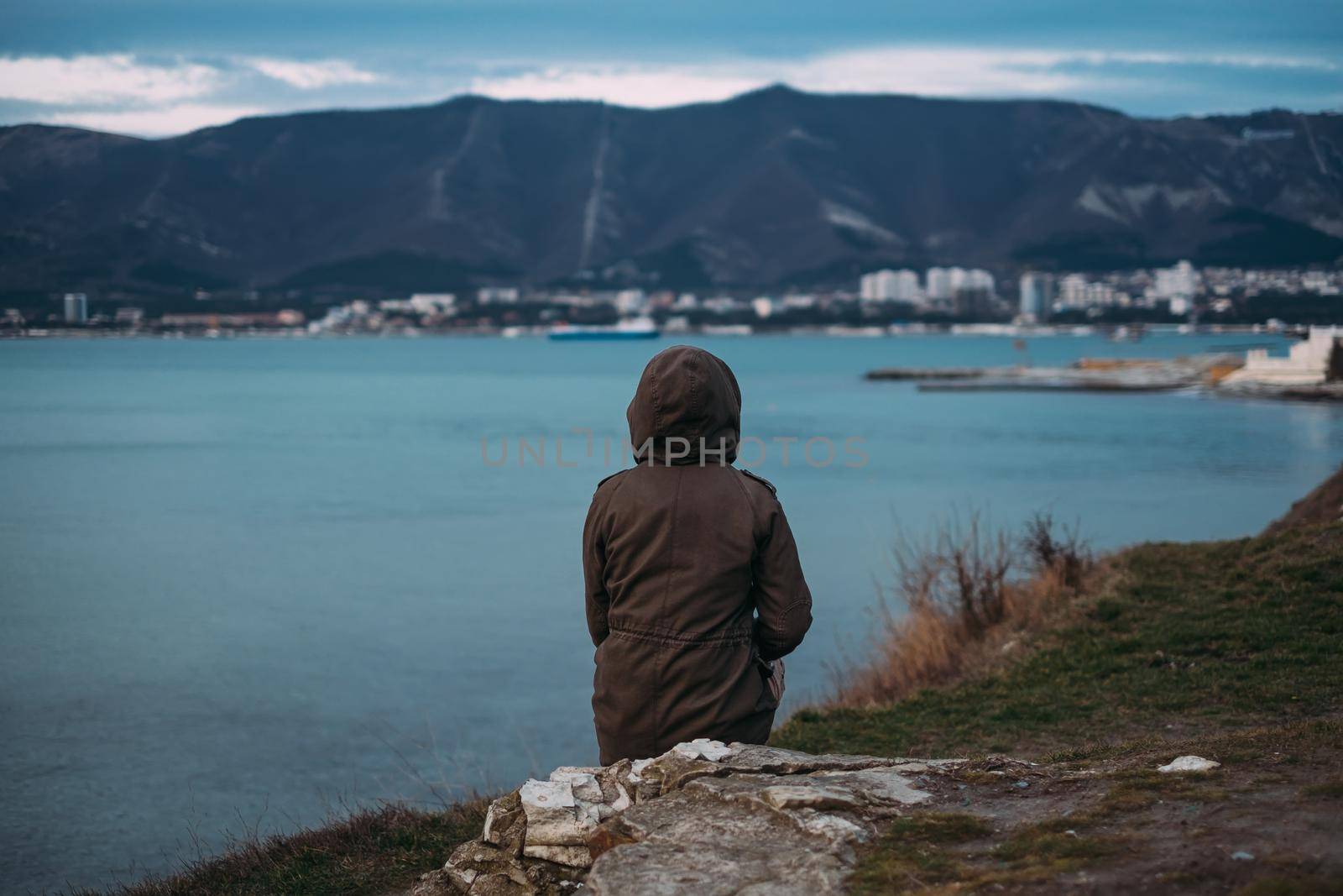 Unrecognizable woman sitting on coast in parka jacket with hood and enjoying view of mountain and sea bay in evening, rear view