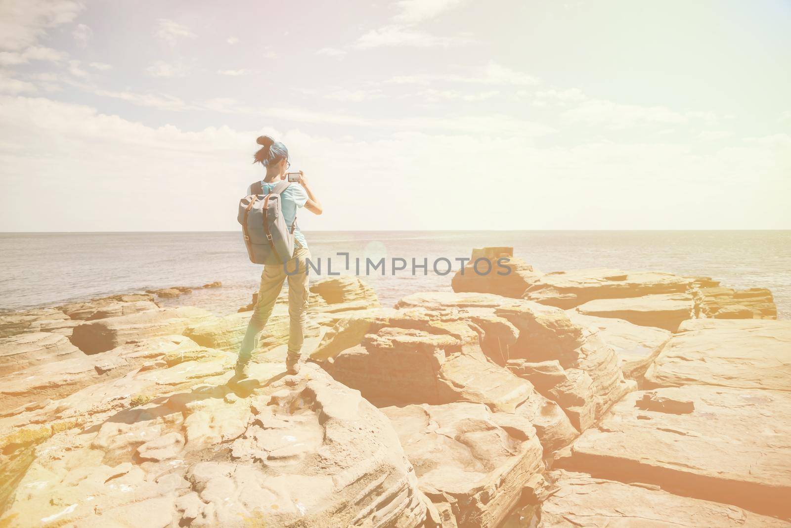 Traveler girl taking photographs beautiful seascape with smartphone on rocky coast in summer. Image with sunlight effect
