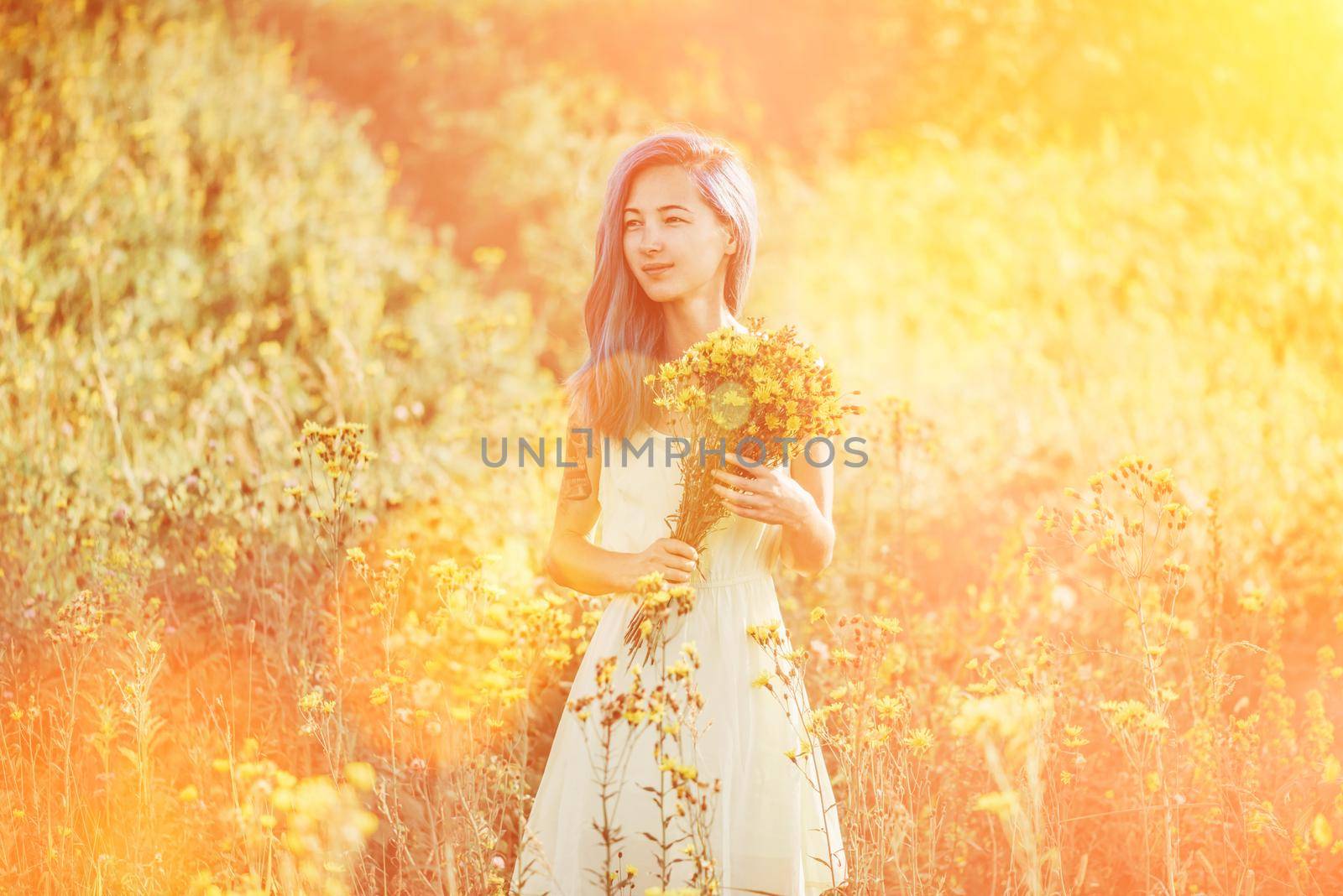 Beautiful young woman with blue hair and bouquet walking on flower field in summer. Image with sunlight effect