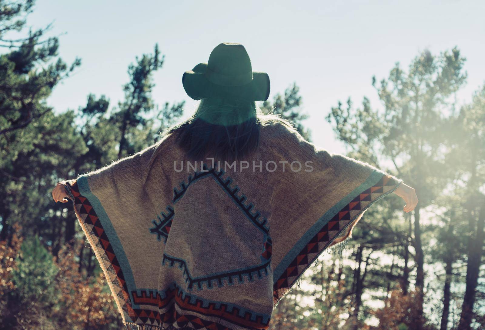 Happy fashionable young woman wearing in poncho and hat standing with raised arms in summer pine forest and enjoying a nice day, rear view