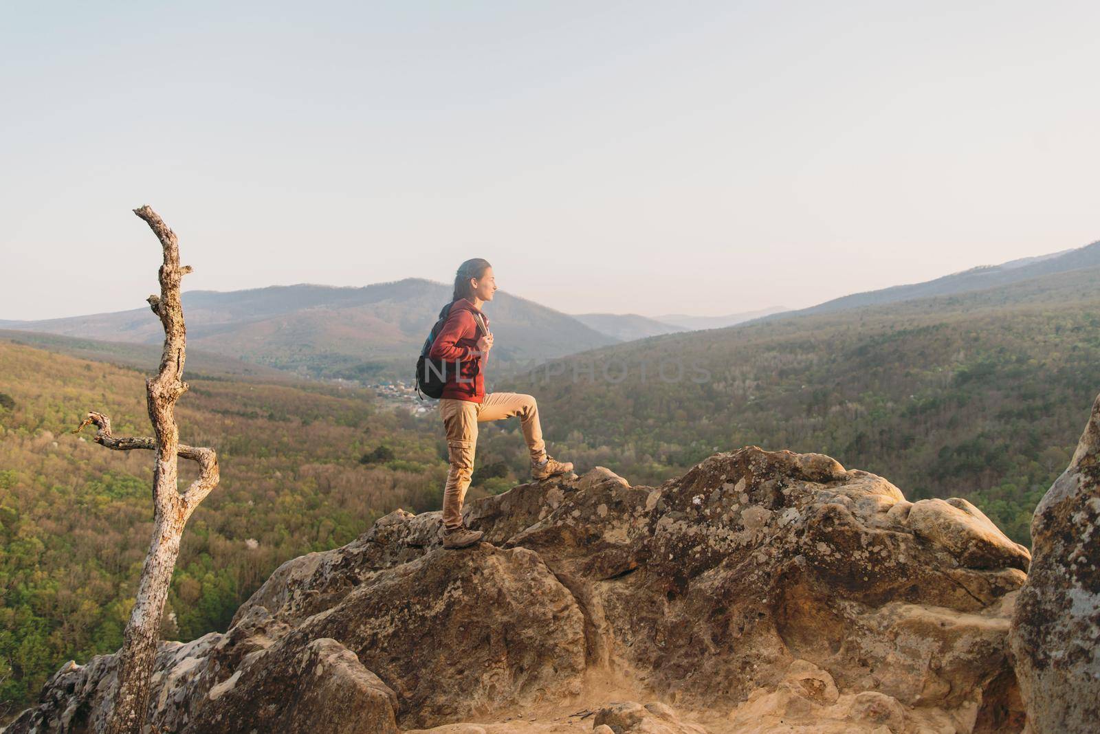 Hiker young woman with backpack standing on peak of rock on background of mountains and looking into the distance in summer outdoor