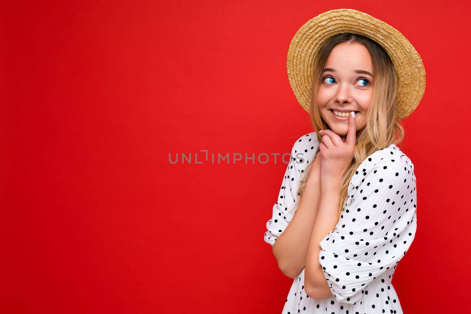 Young beautiful woman. Trendy woman in casual summer dress and straw hat. Positive female shows facial emotions. Funny model isolated on red background with free space by TRMK