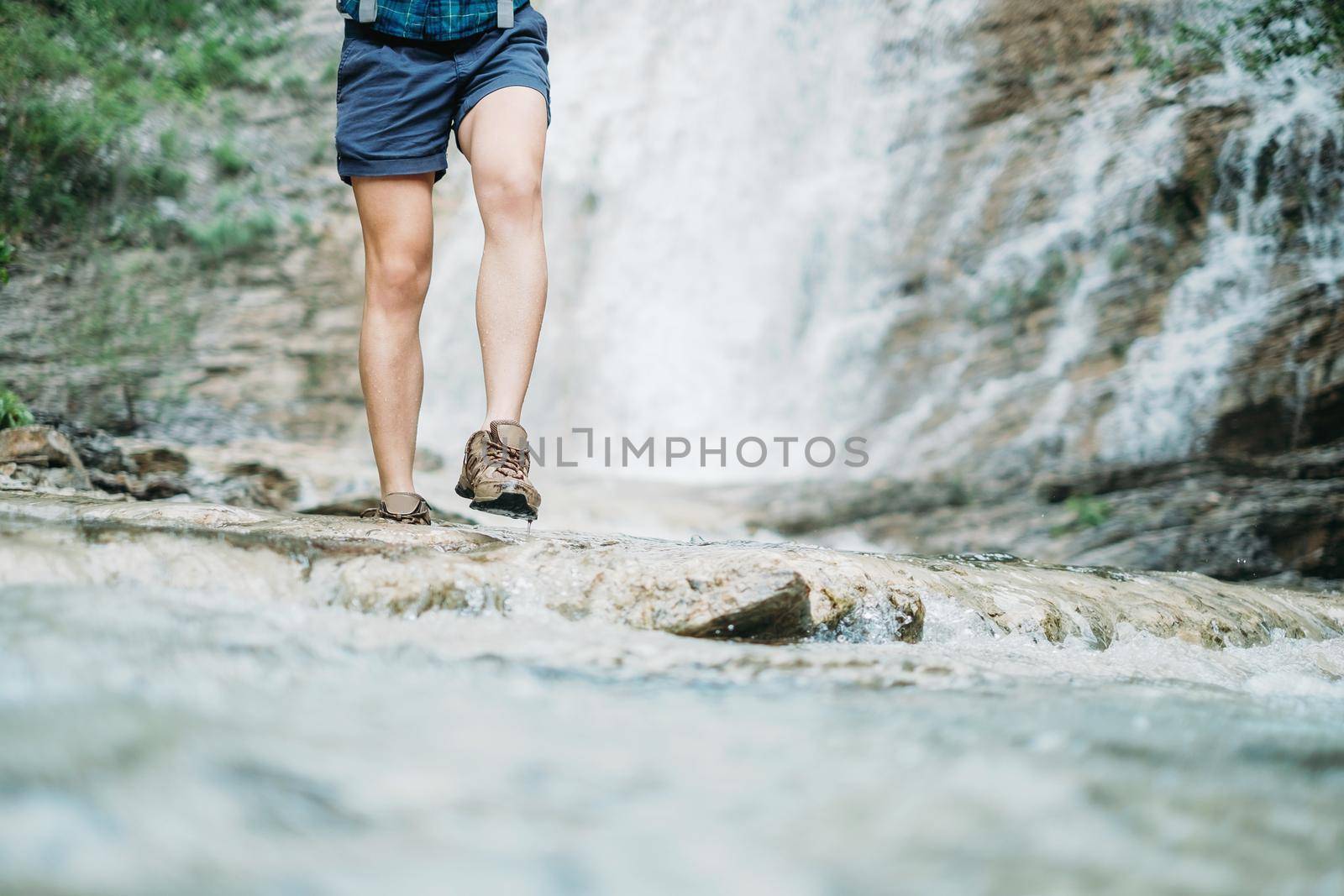Unrecognizable hiker young woman walking along the river on background of waterfall in summer outdoor, view of legs