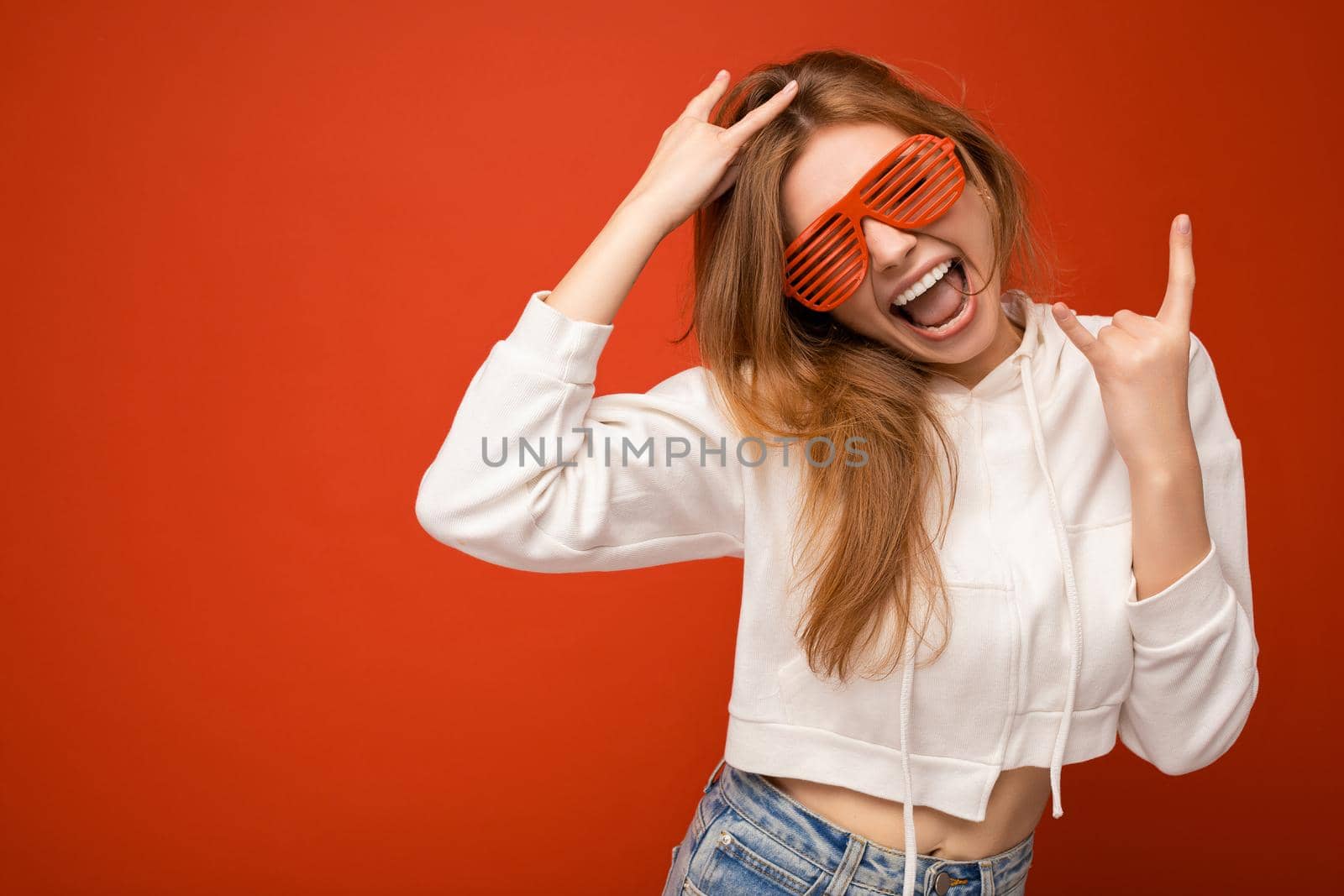 Shot of beautiful emotional positive young blonde woman isolated over colourful background wearing casual white hoodie and stylish sunglasses having fun. empty space