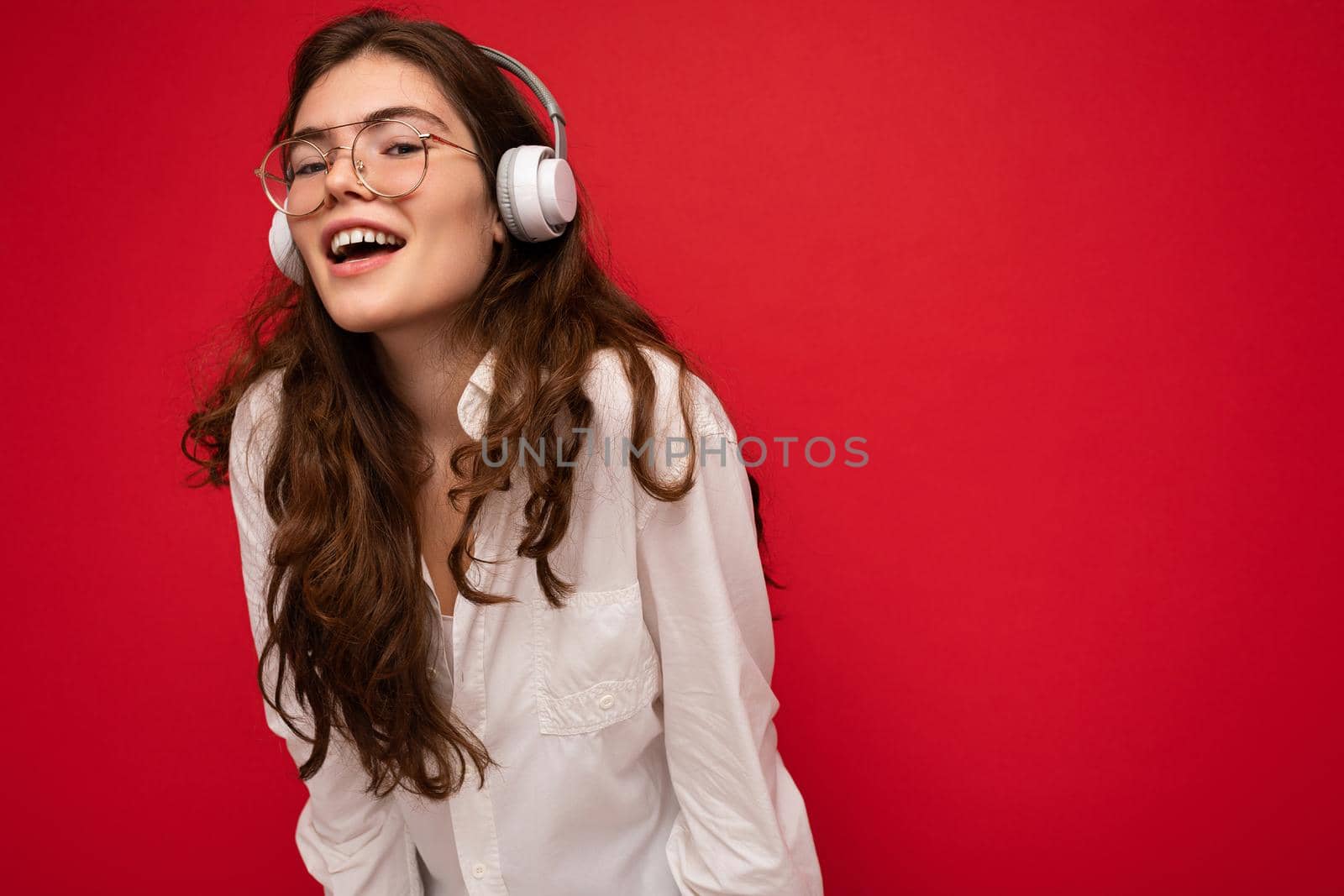 Photo of attractive positive smiling young brunet woman wearing white shirt and optical glasses isolated over red background wearing white wireless bluetooth headsets listening to music and having fun by TRMK