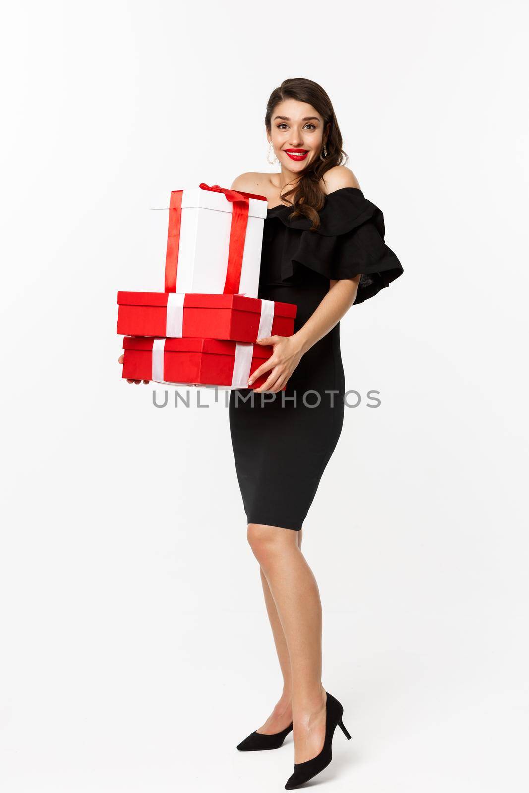 Merry christmas and new year holidays concept. Excited young woman bring gifts, holding xmas presents and smiling at camera, wearing black dress, white background.