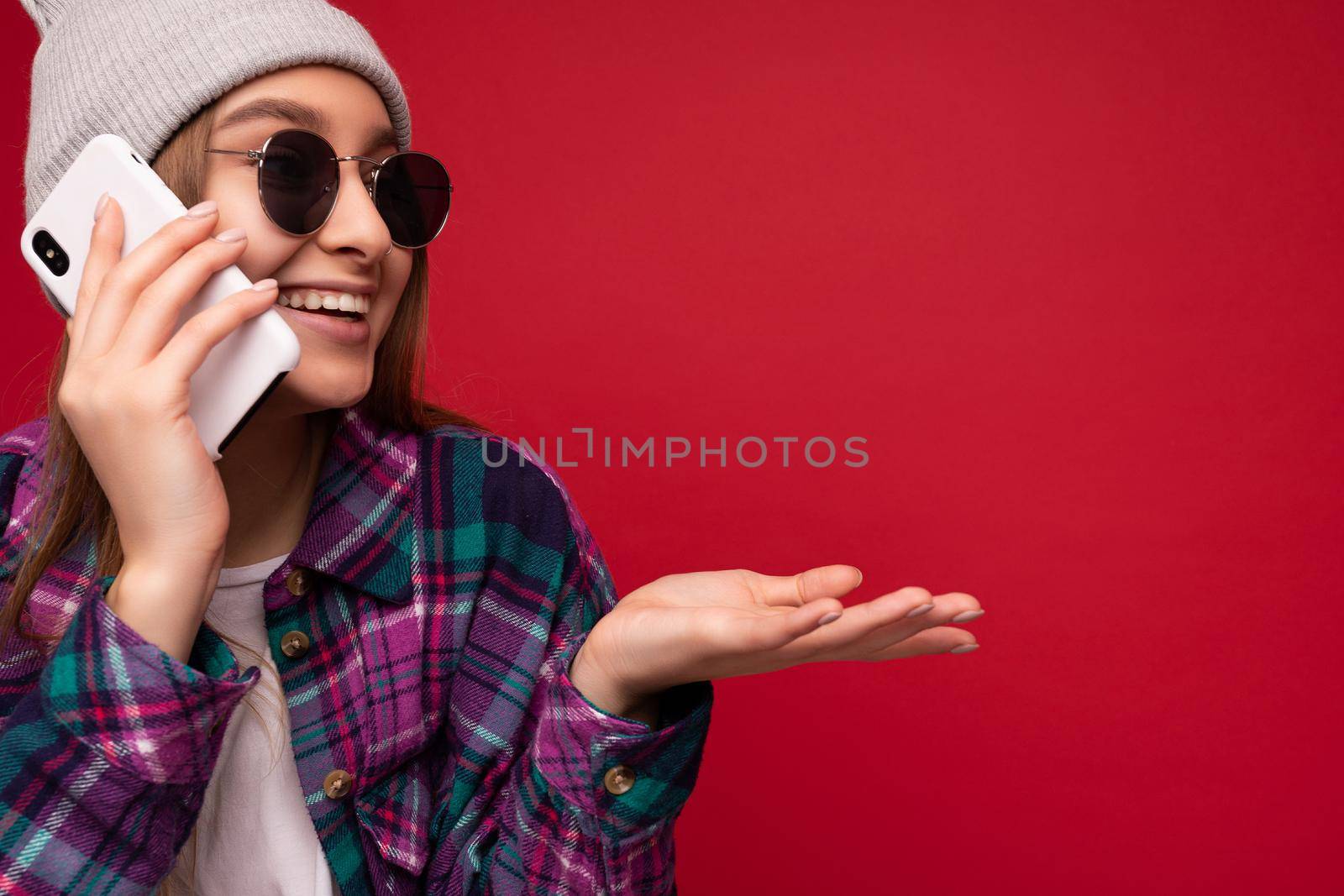 Closeup photo of beautiful emotional positive young blonde woman wearing hipster purple shirt and casual white t-shirt grey hat and sunglasses isolated over red background holding in hand and talking on mobile phone looking to the side by TRMK