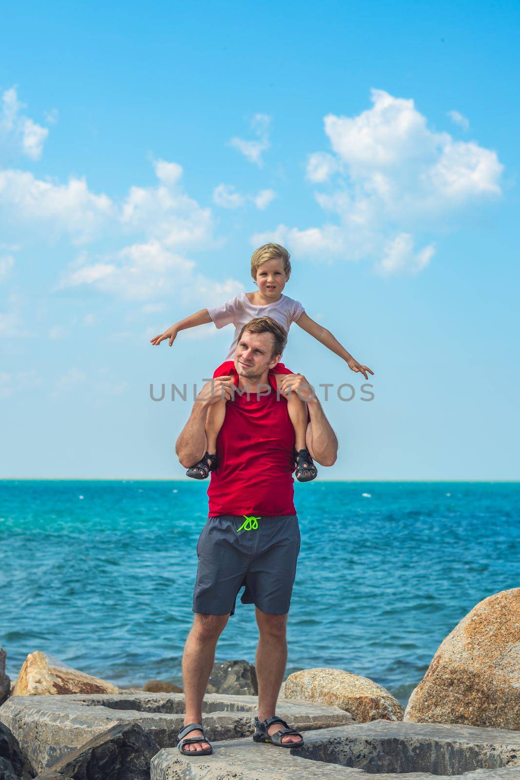 Happy smile father hugs holds on shoulders son boy male child stand blue lazur sea panorama skyline horizon in sunshine day clear sky, play show outstretched hands like airplane. Travel, relationship by nandrey85