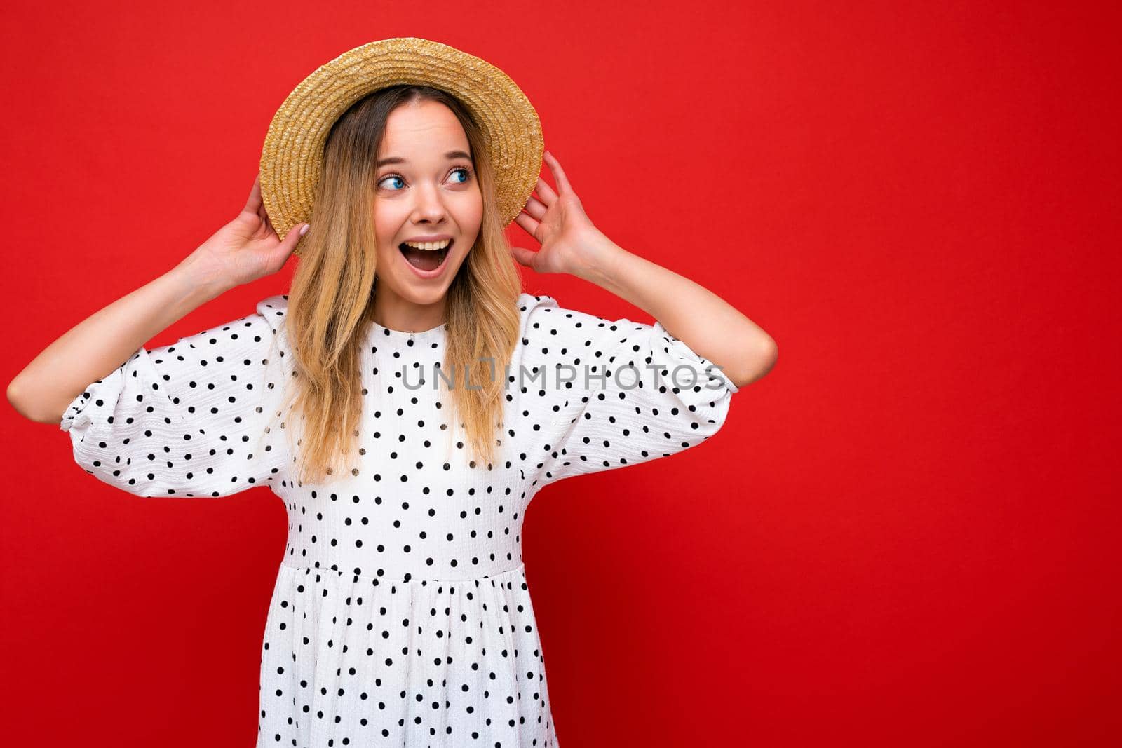 Photo shot of young beautiful cute happy blonde woman wearing casual dress and straw hat isolated over red background with copy space. Summer concept.