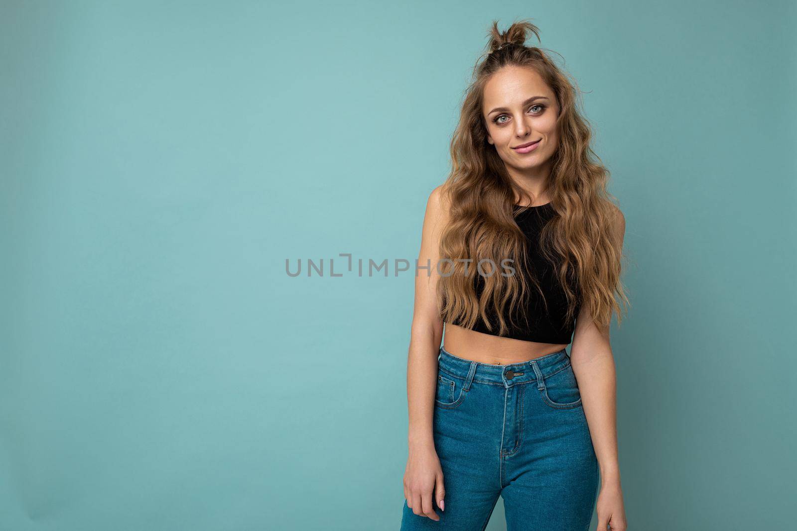 Young beautiful curly blonde woman with sexy expression, cheerful and happy face wearing trendy black top isolated over blue background with copy space.