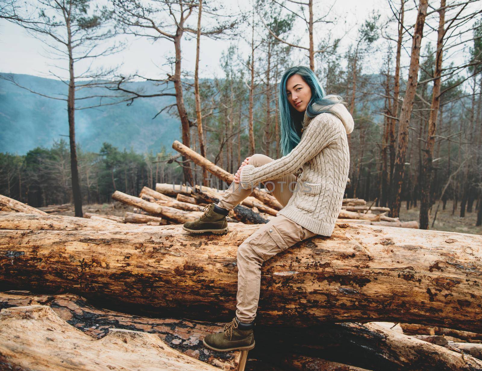 Beautiful young woman sitting on stack of felled tree trunks in the forest and looking at camera