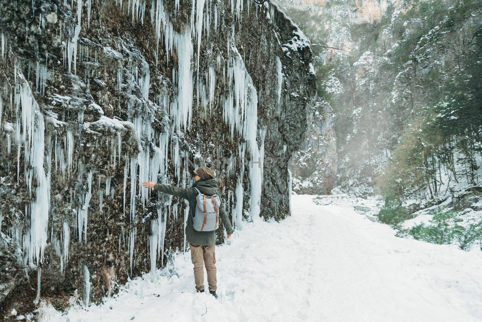 Hiker young woman with backpack walking in gorge and touching wall of icicles in winter