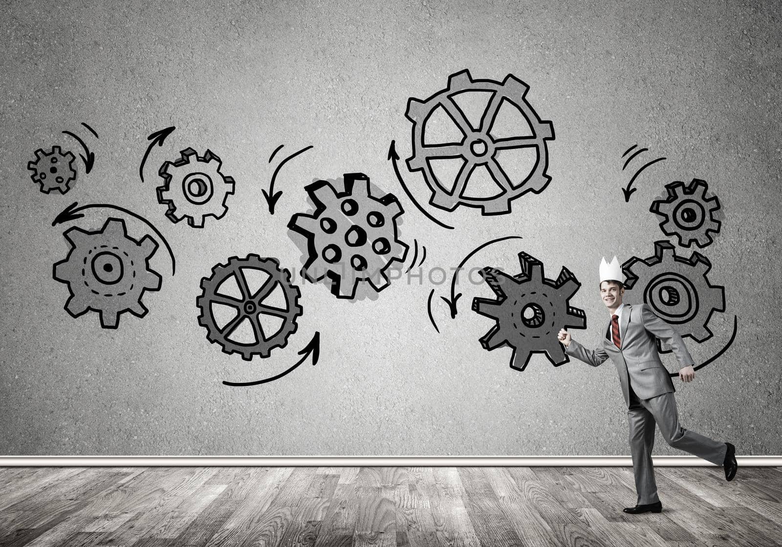 Young handsome businessman wearing white paper crown running and gear mechanism on wall