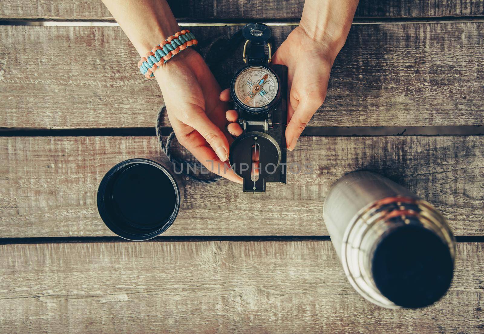 Traveler woman holding compass on wooden table near the thermos, point of view. Toned image