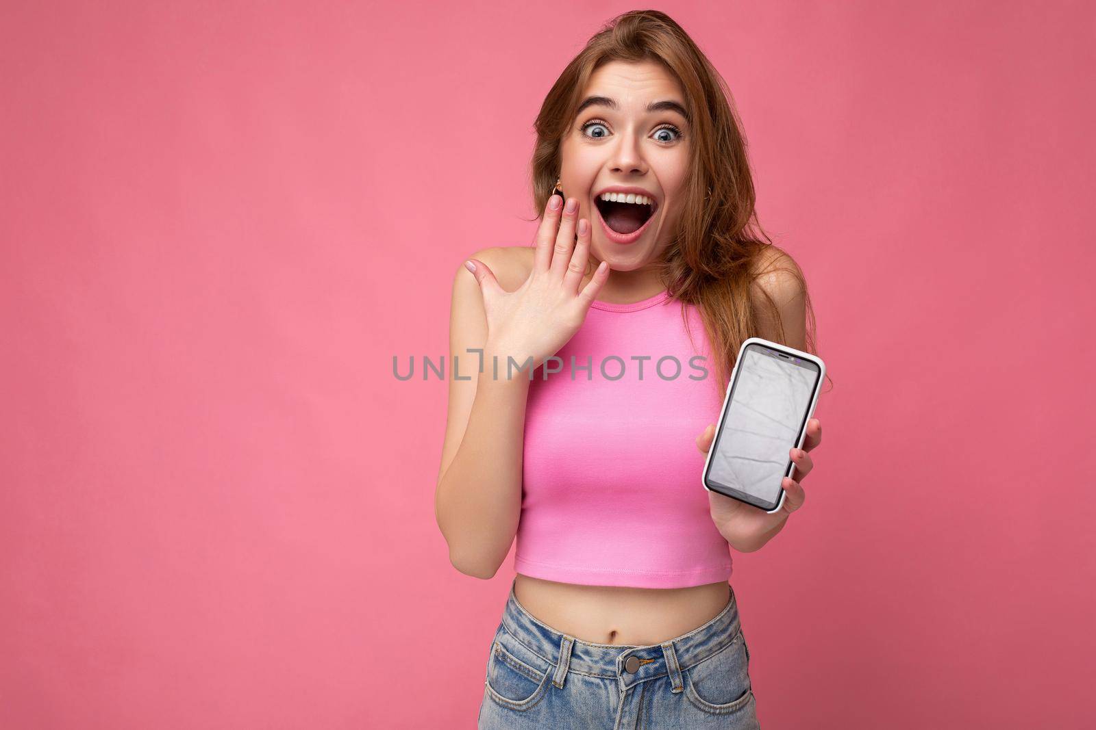 Photo of beautiful surprised amazed smiling young woman good looking wearing casual stylish outfit standing isolated on background with copy space holding smartphone showing phone in hand with empty screen display for mockup pointing at gadjet looking at camera by TRMK