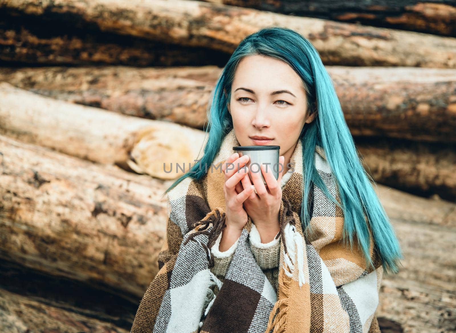 Young woman wrapped in plaid sitting with cup of tea on background of tree trunks outdoor. Toned image