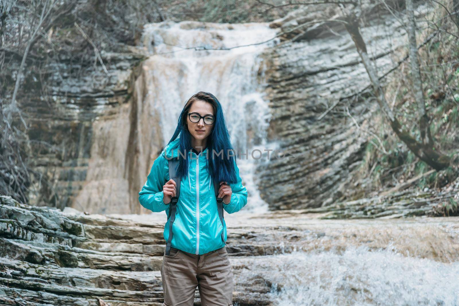 Beautiful traveler young woman with backpack standing on background of waterfall and looking at camera outdoor