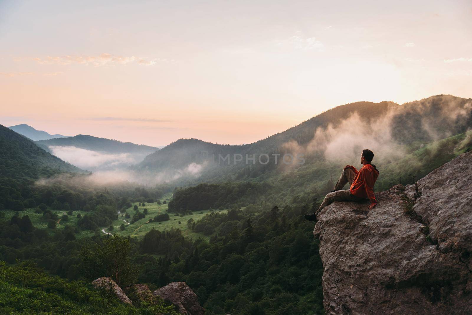 Traveler young man sitting on rocky stone and enjoying view of sunset in the mountains