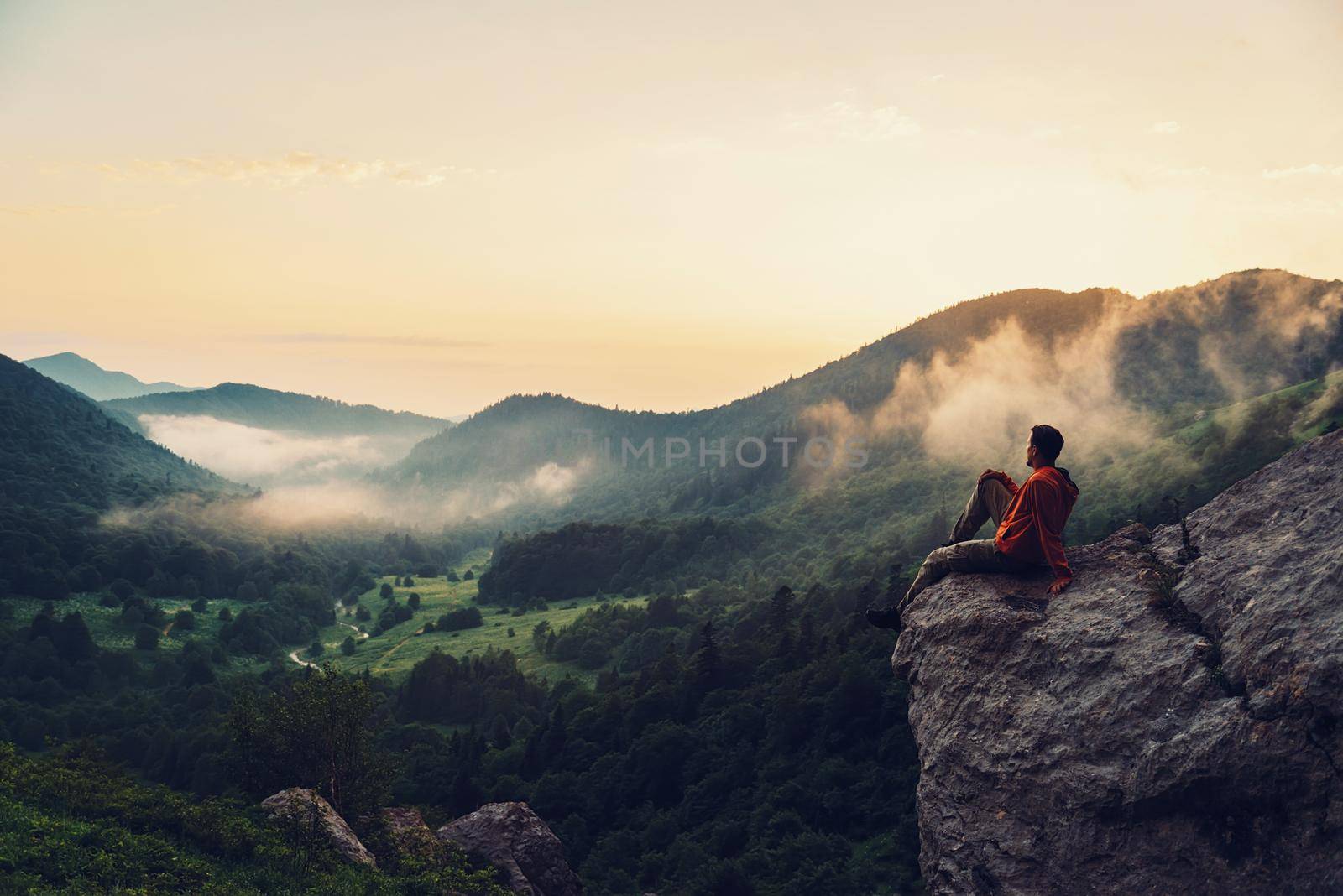 Traveler young man sitting on rocky stone and enjoying view of sunset in the mountains. Toned image