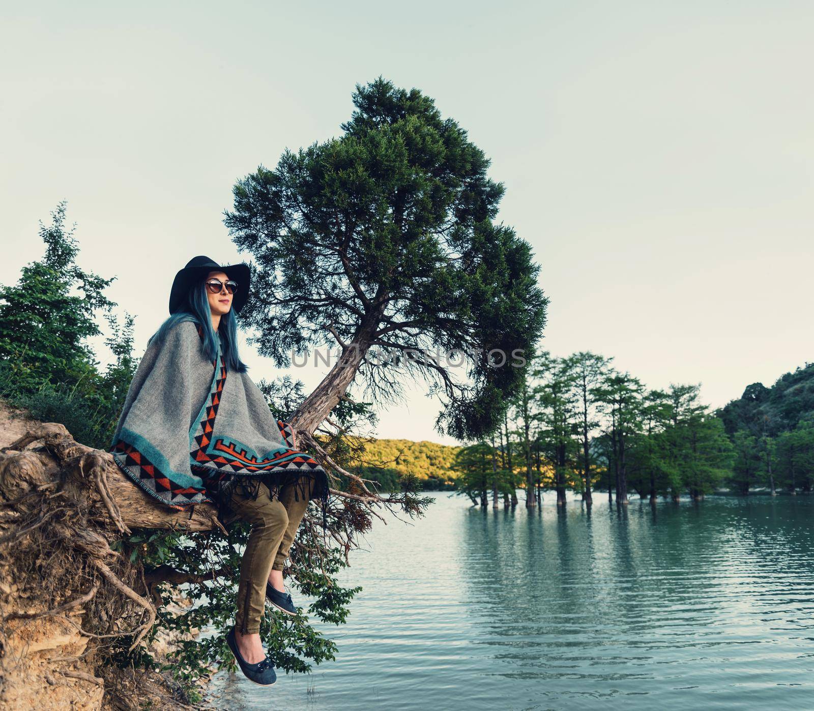 Smiling beautiful young woman resting on tree on the shore of lake in summer