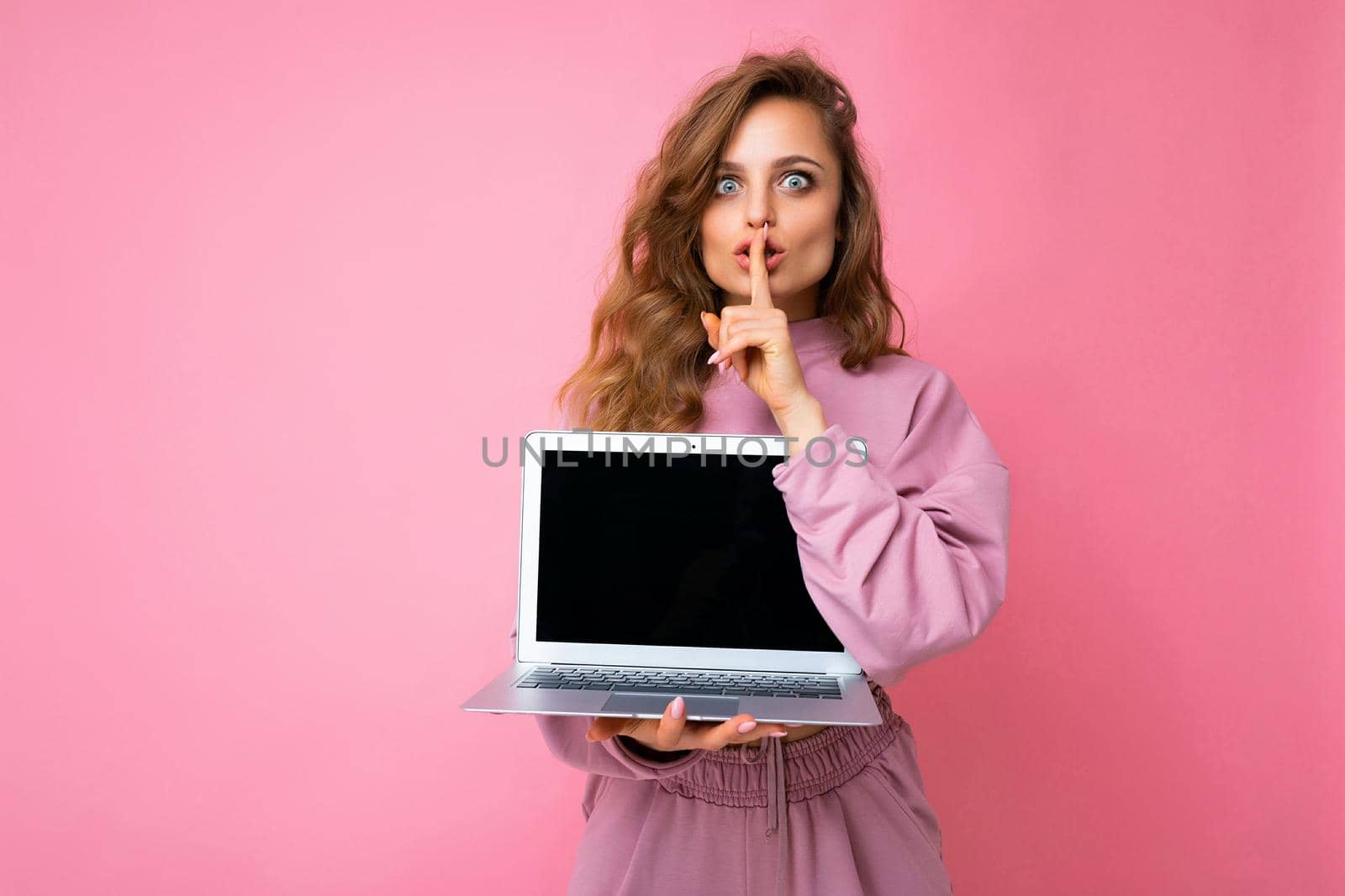 Close-up portrait of beautiful dark blond curly young woman holding laptop computer looking at camera holding finger close to mouth and lips showing gesture shhh and saying hush be quiet wearing pink sweater isolated over pink wall background. Mock up copy empty space..