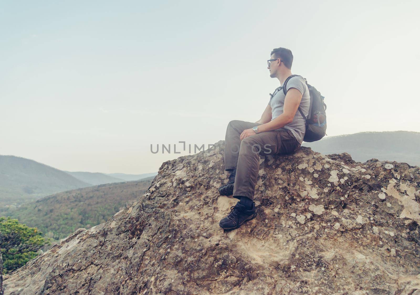 Hiker young man with backpack sitting on peak of rock on background of mountains and enjoying view of landscape in summer outdoor