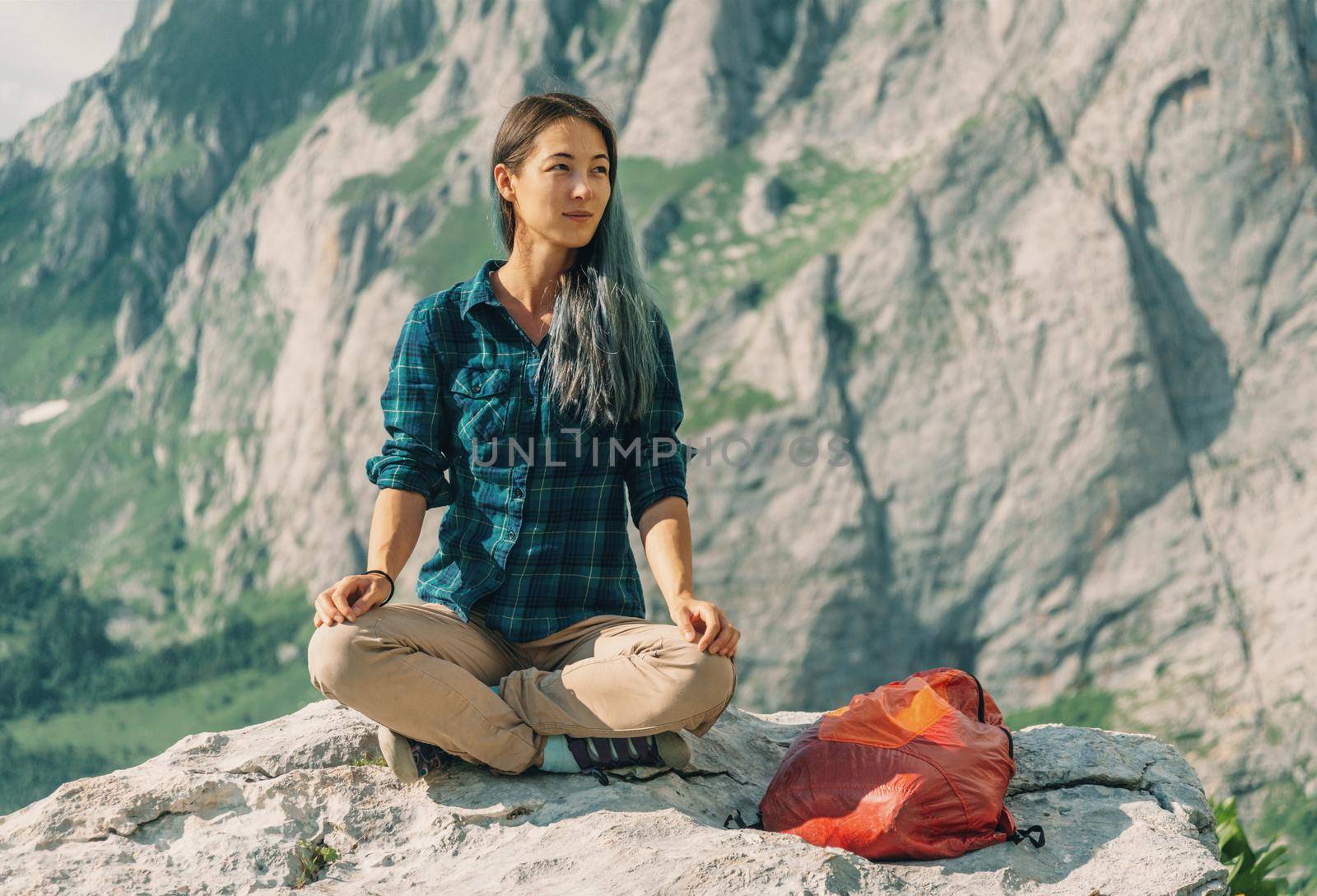 Smiling female hiker sitting in pose of lotos on stone in summer on background of cliffs outdoor