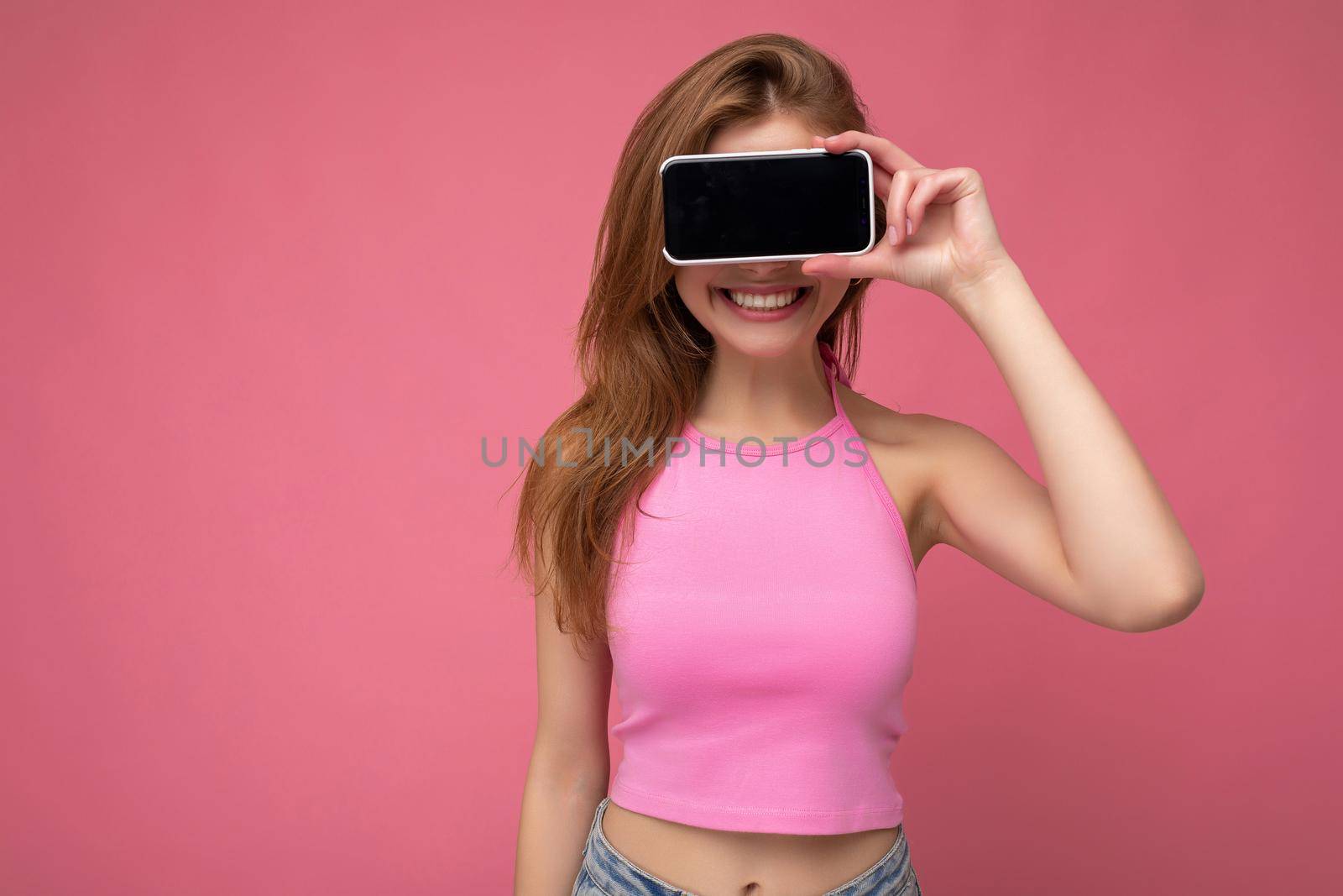 Photo of beautiful smiling young woman good looking wearing casual stylish outfit standing isolated on background with copy space holding smartphone showing phone in hand with empty screen display for mockup.