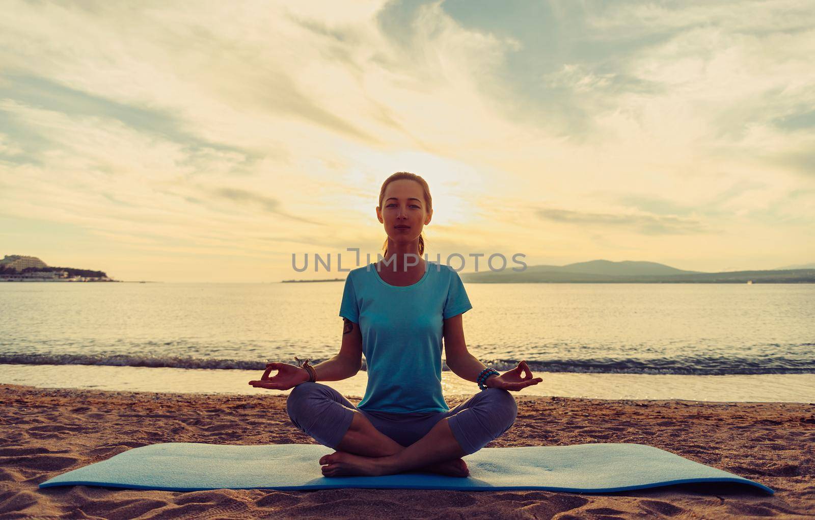 Young woman meditating in pose of lotus on sand beach near the sea at sunset in summer, front view