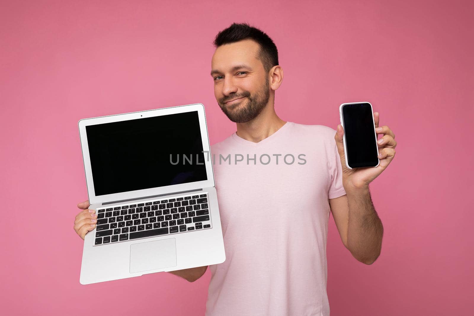 Handsome brunet man holding laptop computer and mobile phone looking at camera in t-shirt on isolated pink background.