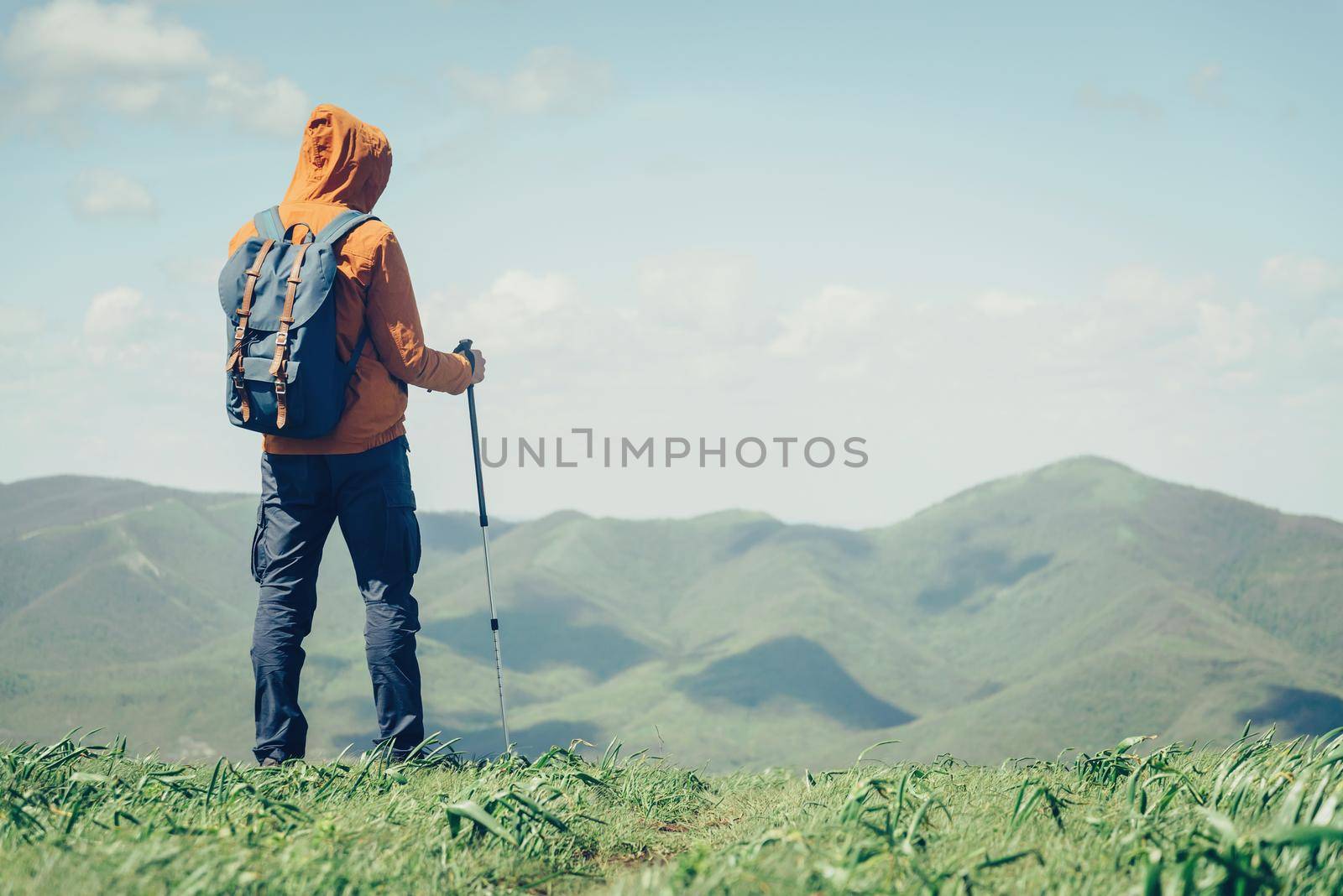 Unrecognizable traveler young man with backpack and trekking poles looking at mountains in summer outdoor. Free space in right part of the image