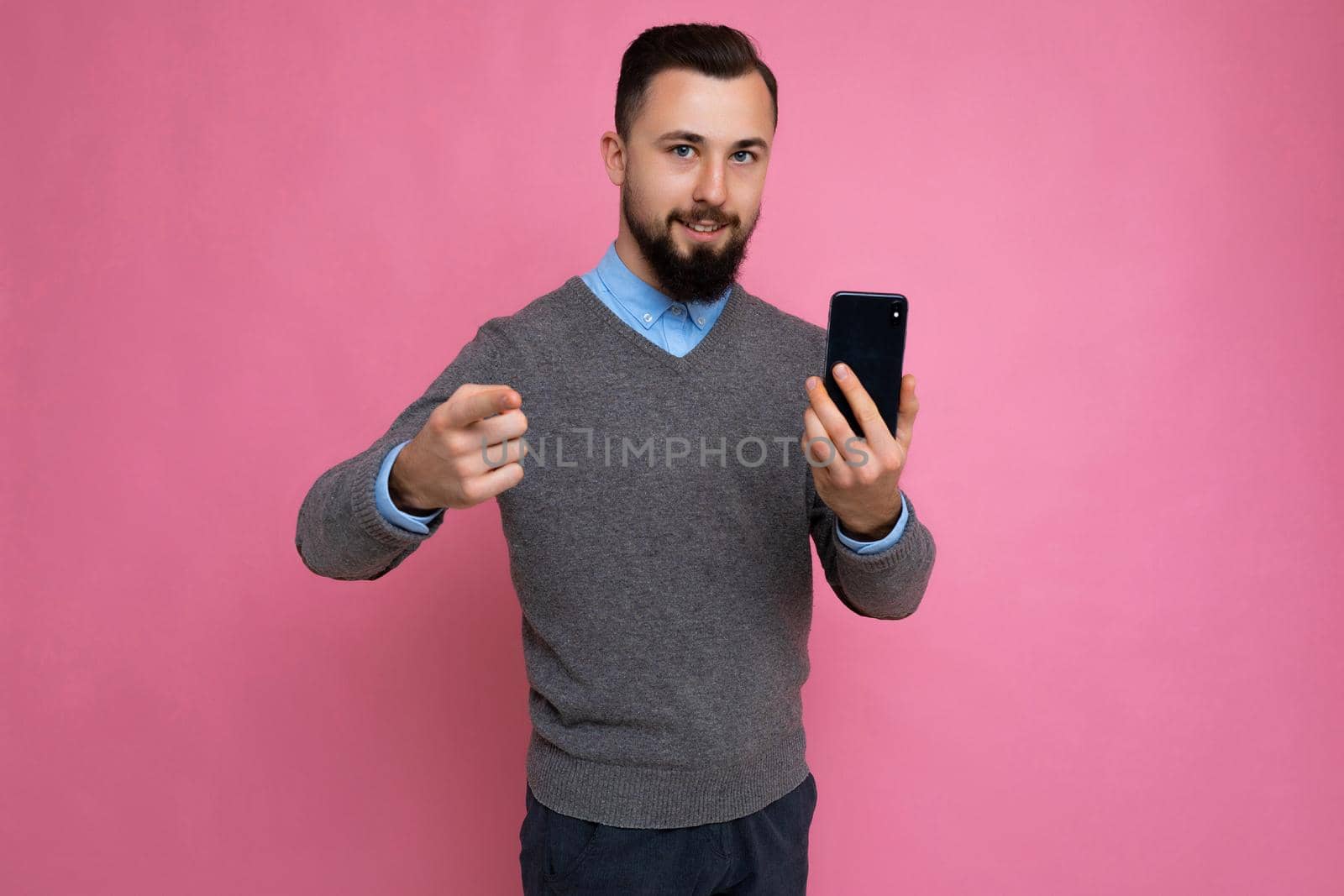 Good looking happy cool young brunette unshaven man with beardwearing stylish grey sweater and blue shirt standing isolated over pink background wall holding smartphone and using phone looking at camera pointing finger at camera.