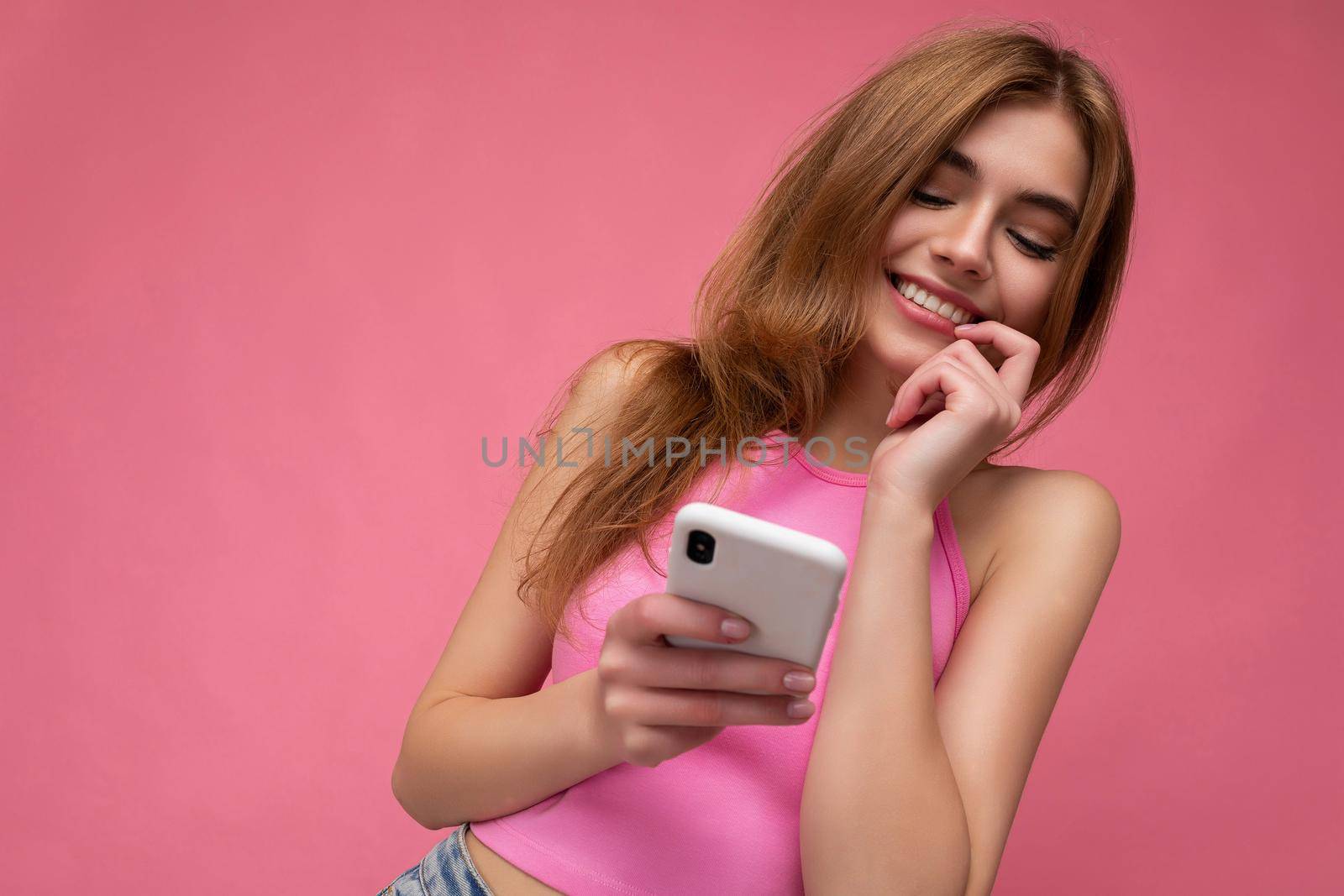 Photo of good looking young blonde woman wearing pink top poising isolated on pink background with empty space holding in hand and using mobile phone messaging sms looking at gadjet display and thinking.