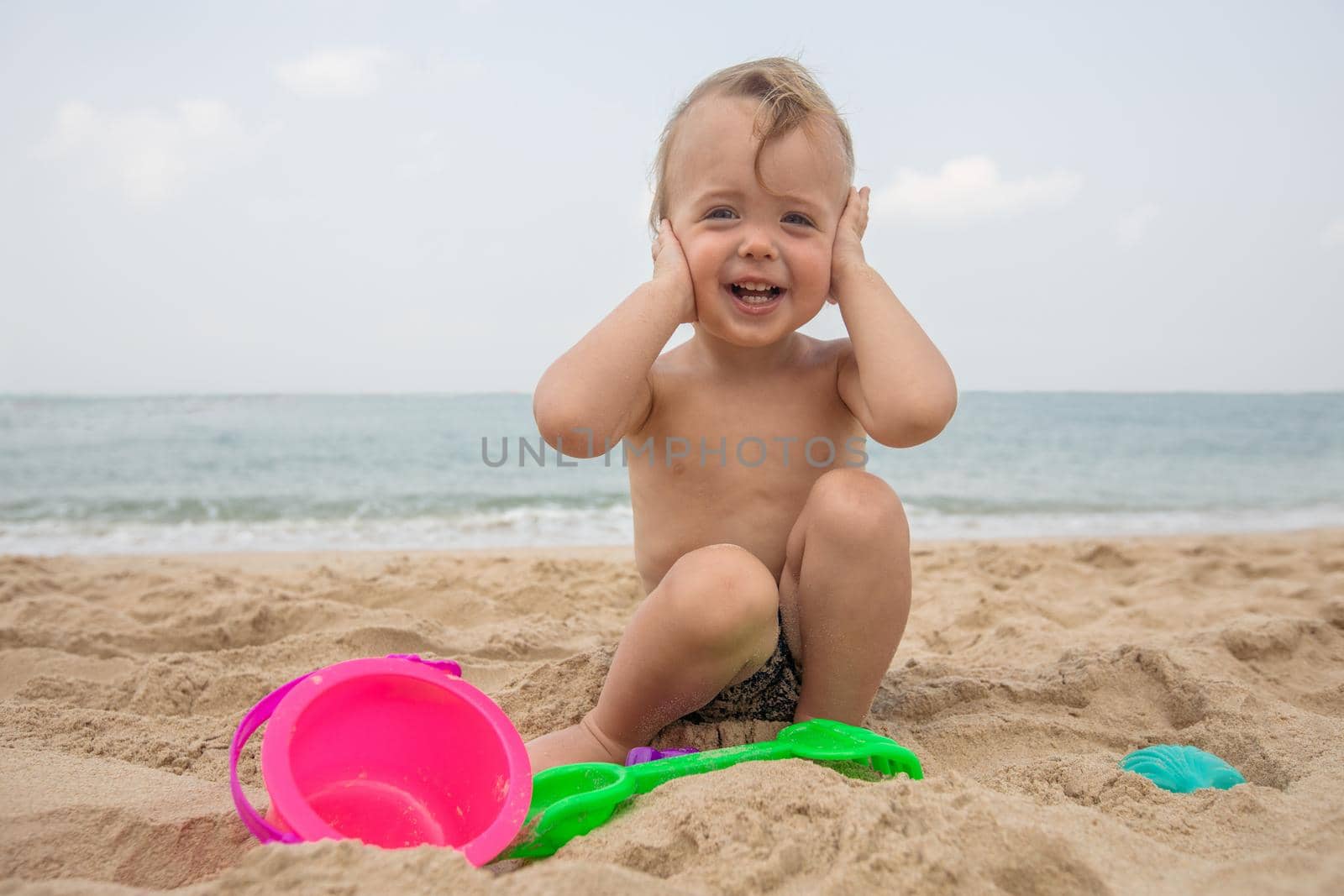 Charming little boy with toys sitting on sand and laughing at camera on seascape.