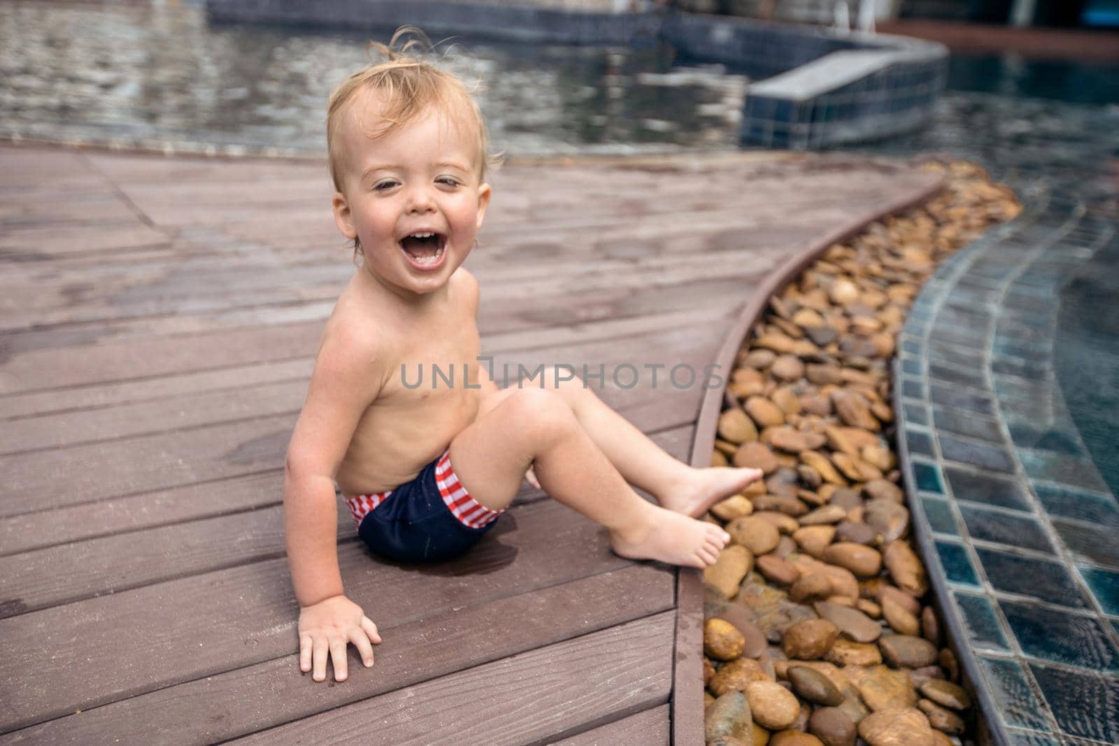 Innocent happy infant sitting on wooden poolside of resort and laughing excitedly.