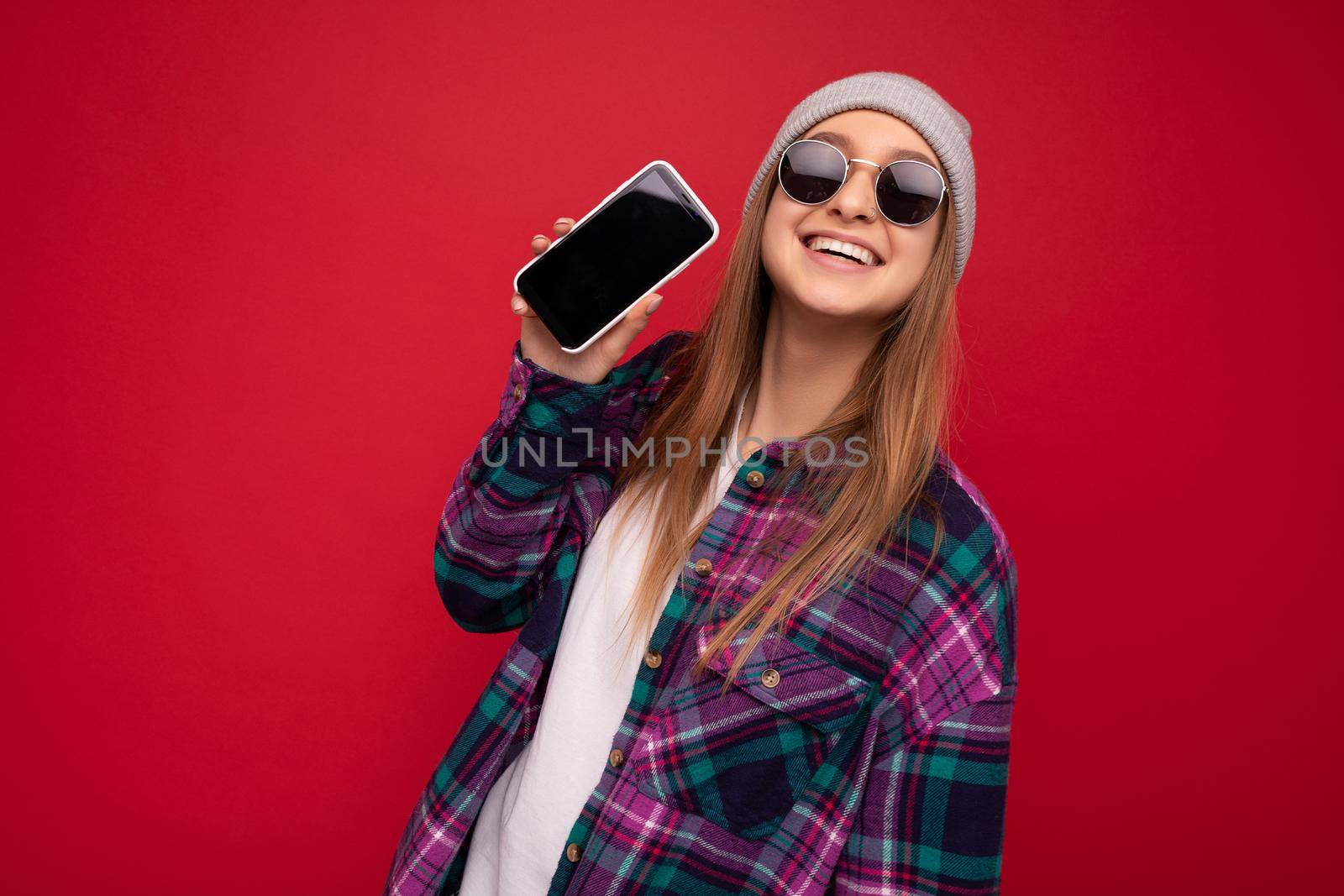 Photo of beautiful positive young blonde woman wearing stylish purple shirt and casual white t-shirt grey hat and sunglasses isolated over red background holding in hand and showing mobile phone with empty screen for mockup looking at camera.