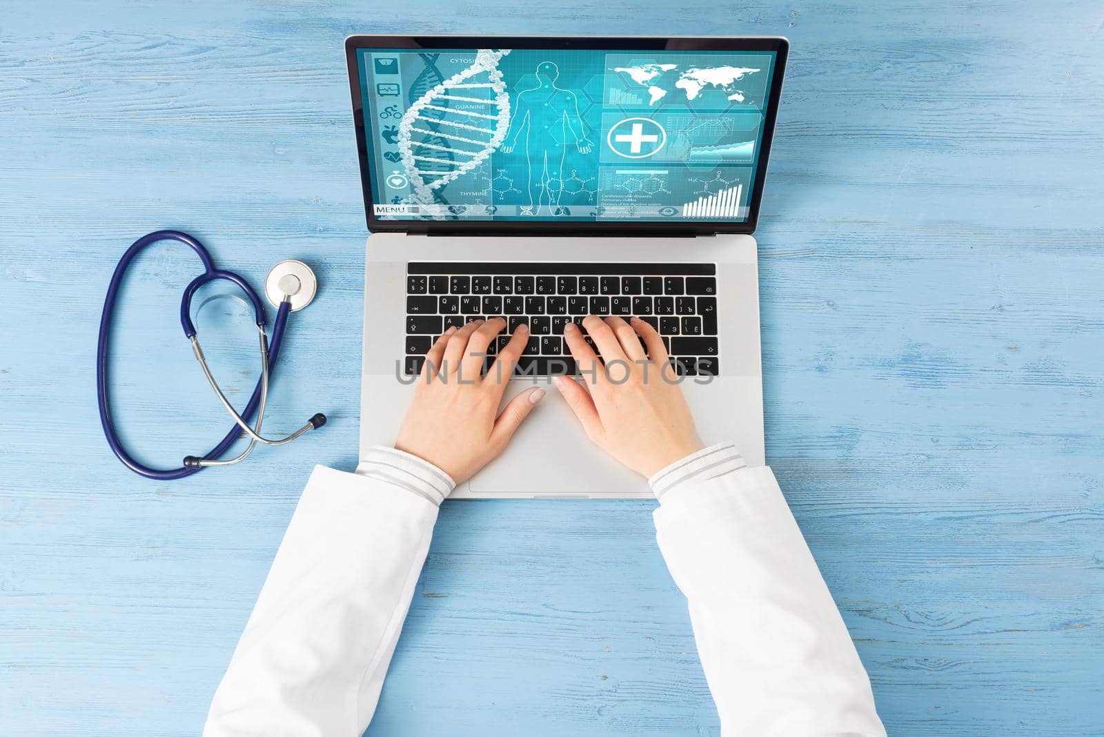 Top view of doctor hands typing at laptop computer. Therapist in white coat sitting at blue wooden desk. Online medical clinic communication with patient. Medical application and healthcare services.