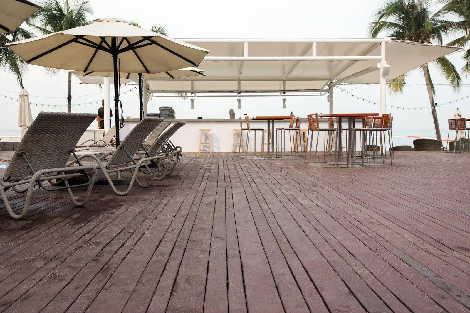 Wooden deck with lounge sunbeds and bar area on background of palms on tropical resort.